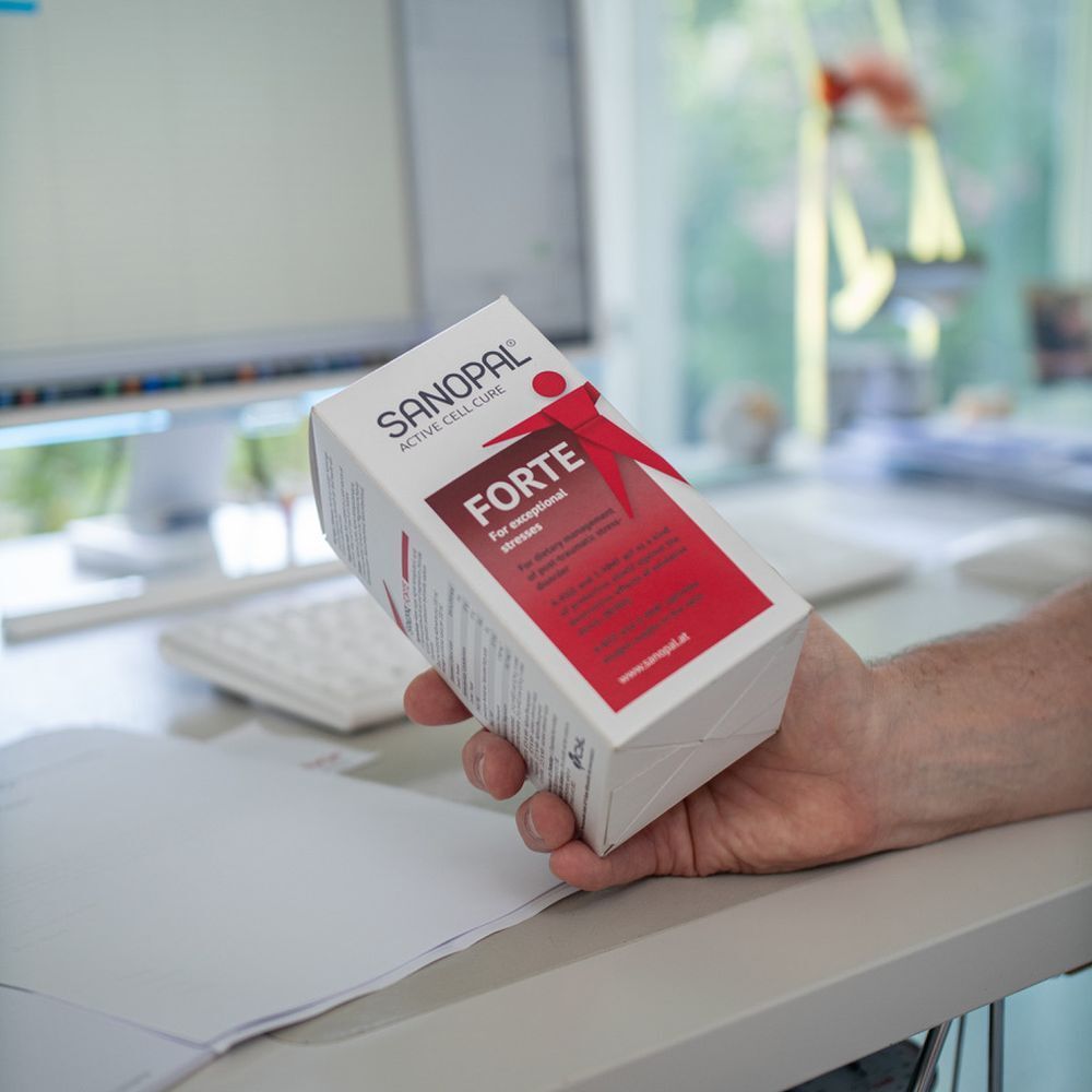 Hand hält Sanopal Forte-Verpackung. Weißer Karton mit roter Schrift und Logo. Auf einem Schreibtisch.