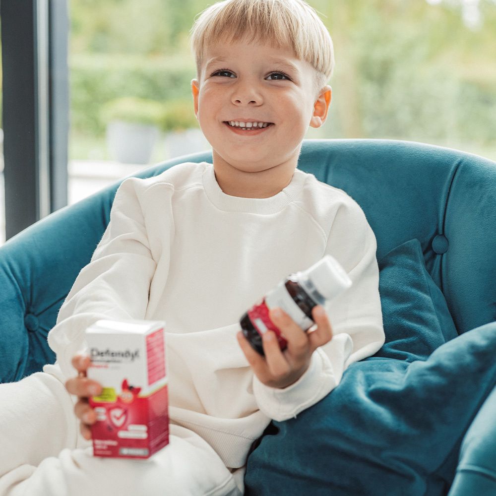 Junge hält Defendyl-Imunoglukan P4H® FORTE Flasche und Verpackung. Kind lächelt. Blauer Sessel im Hintergrund.