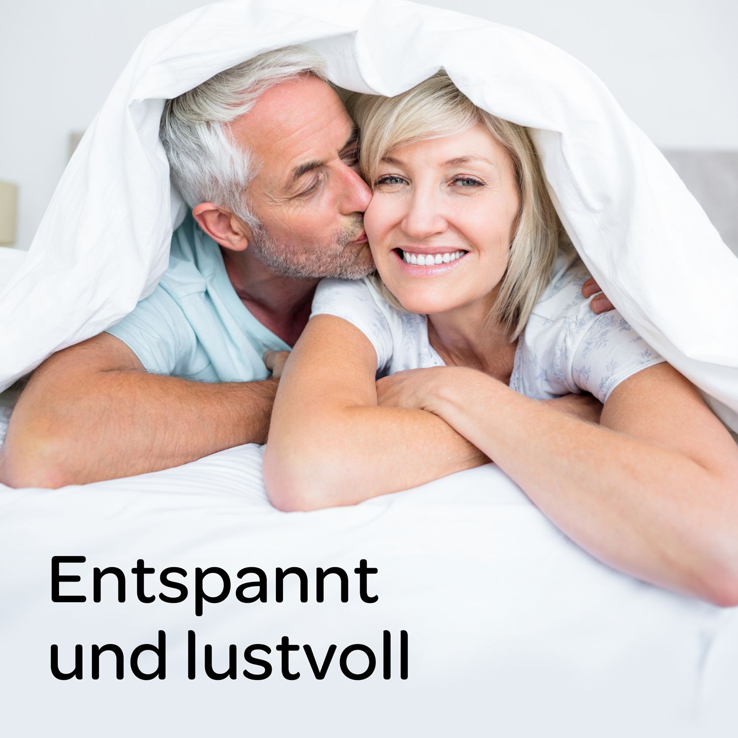 Ein älteres Paar liegt unter einer weißen Decke. Der Mann küsst die Frau auf die Wange. Text: Entspannt und lustvoll.