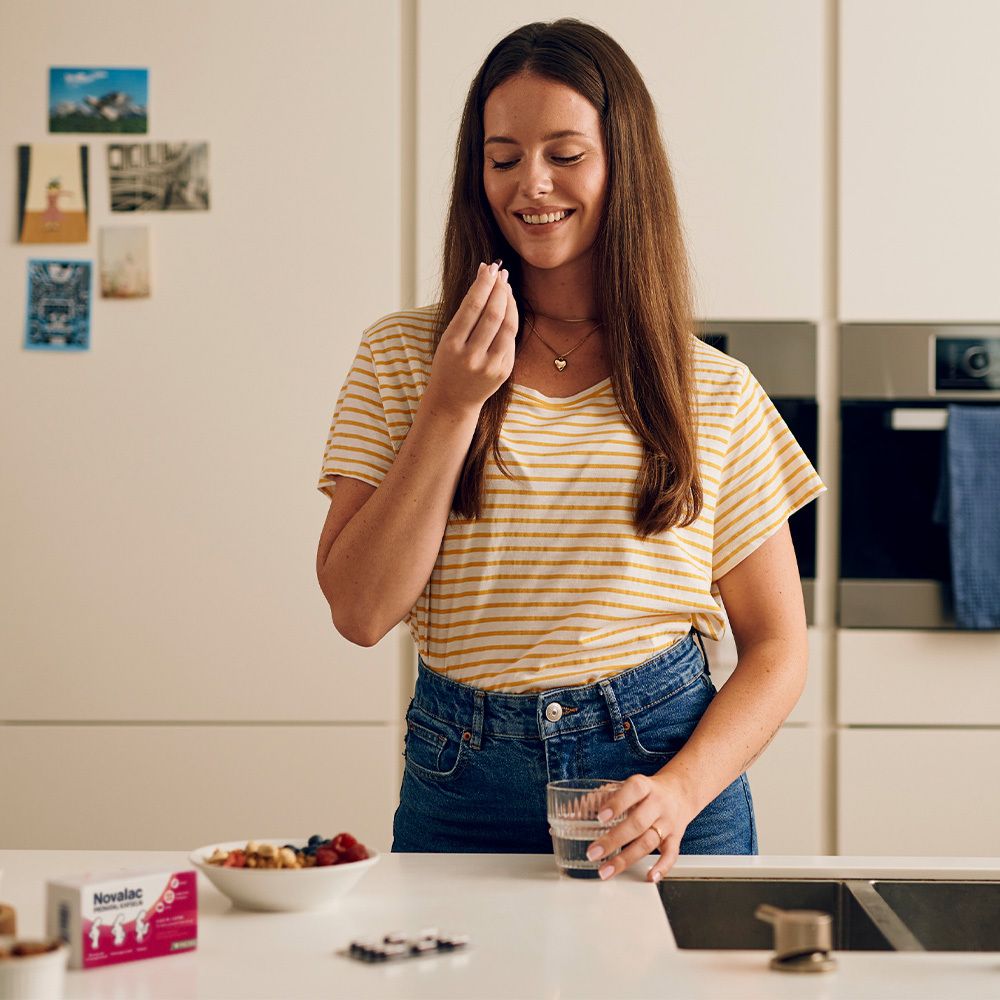 Frau in Küche hält Kapsel. Auf Tisch: Novalac PRENATAL KAPSELN Packung, Obstschale, Glas Wasser. Gelb-weiß gestreiftes T-Shirt.