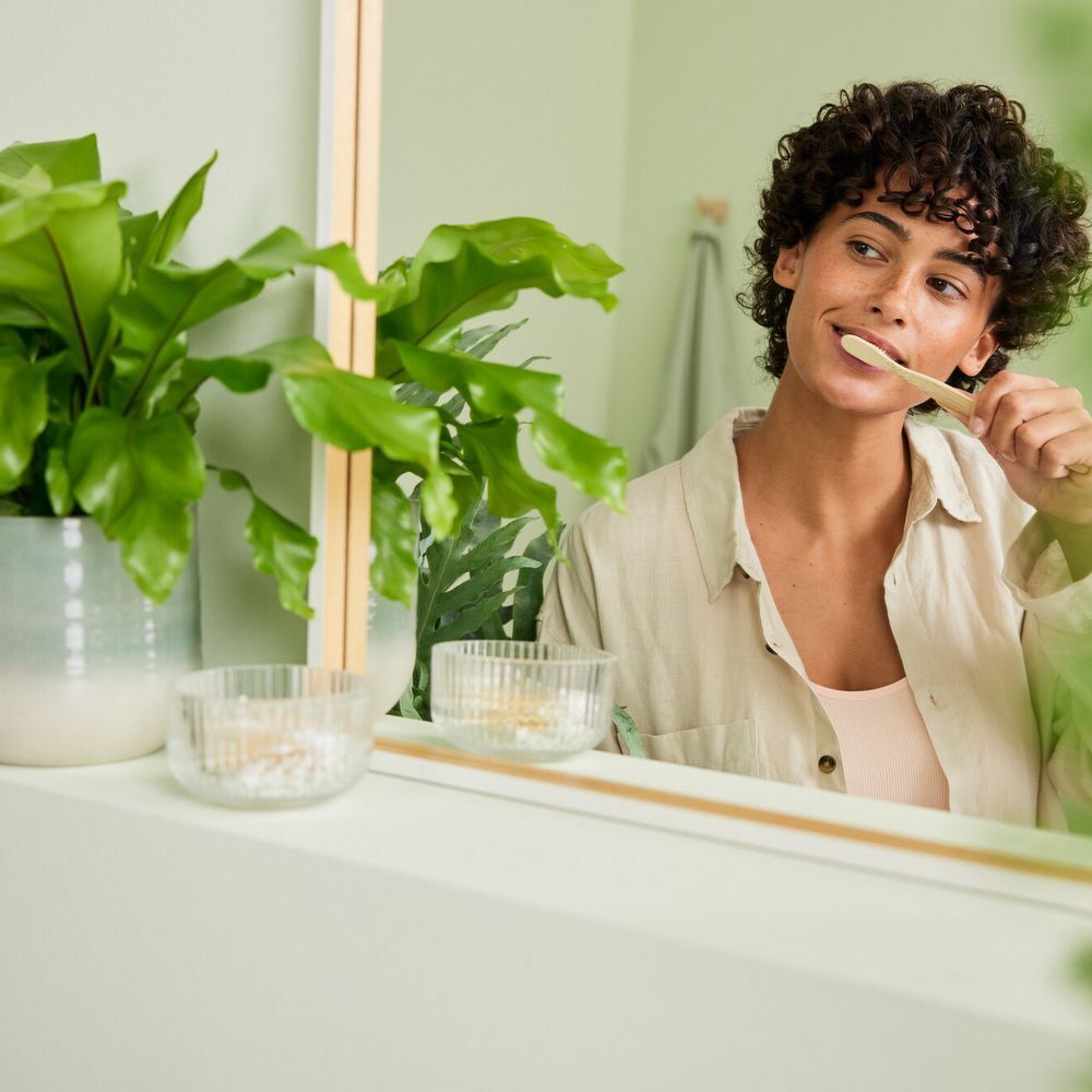 Femme se brossant les dents devant un miroir. Plante en arrière-plan. Deux bols sur le lavabo.