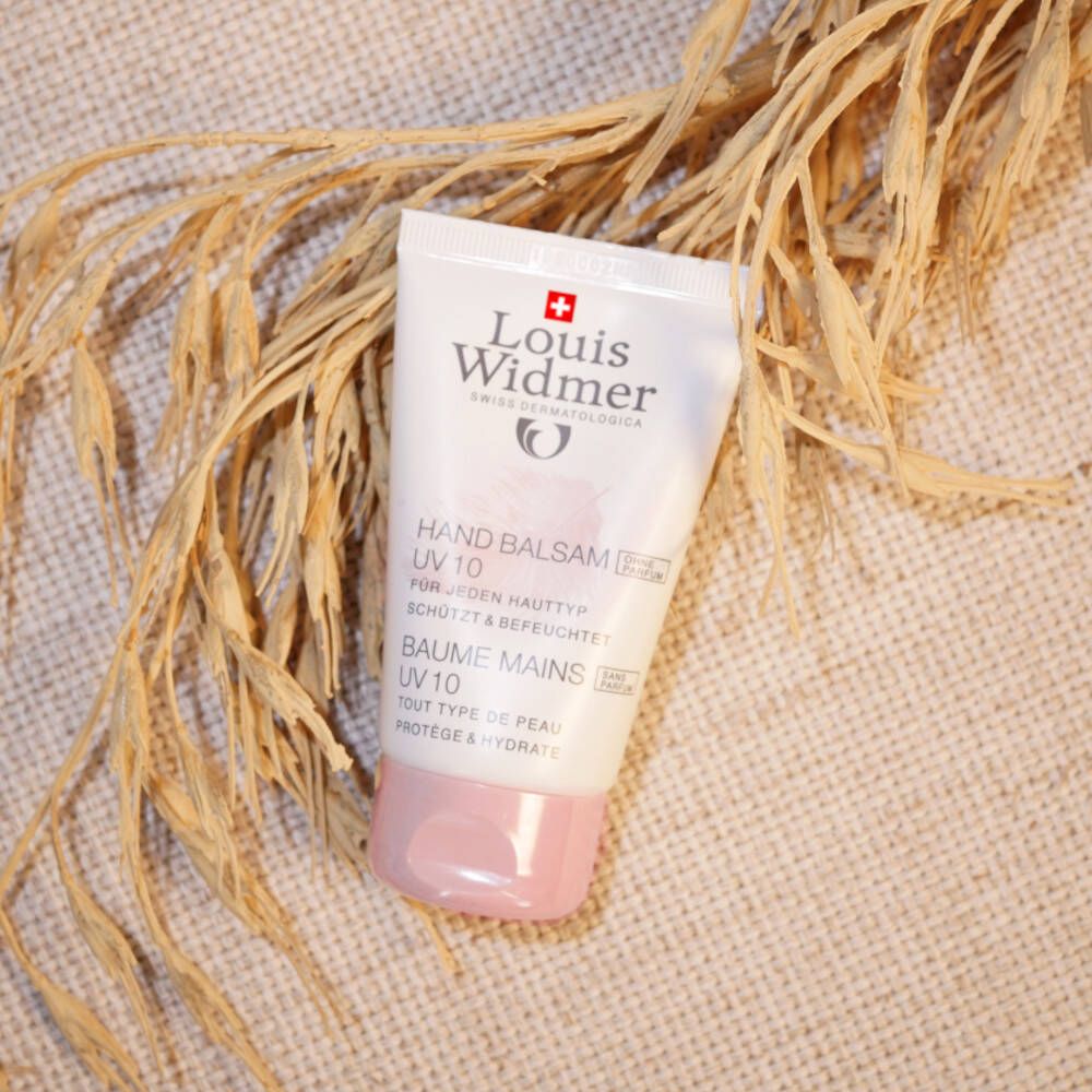 Tube de baume pour les mains blanc avec bouchon rose, à côté d'épis séchés. Inscription: Louis Widmer, Hand Balsam UV 10.