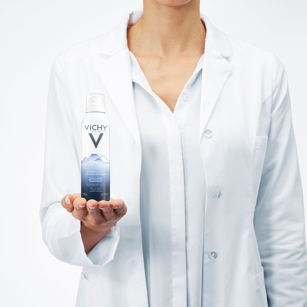Persoon houdt een sprayfles vast. Draagt een witte laboratoriumjas. Productnaam en logo zichtbaar.