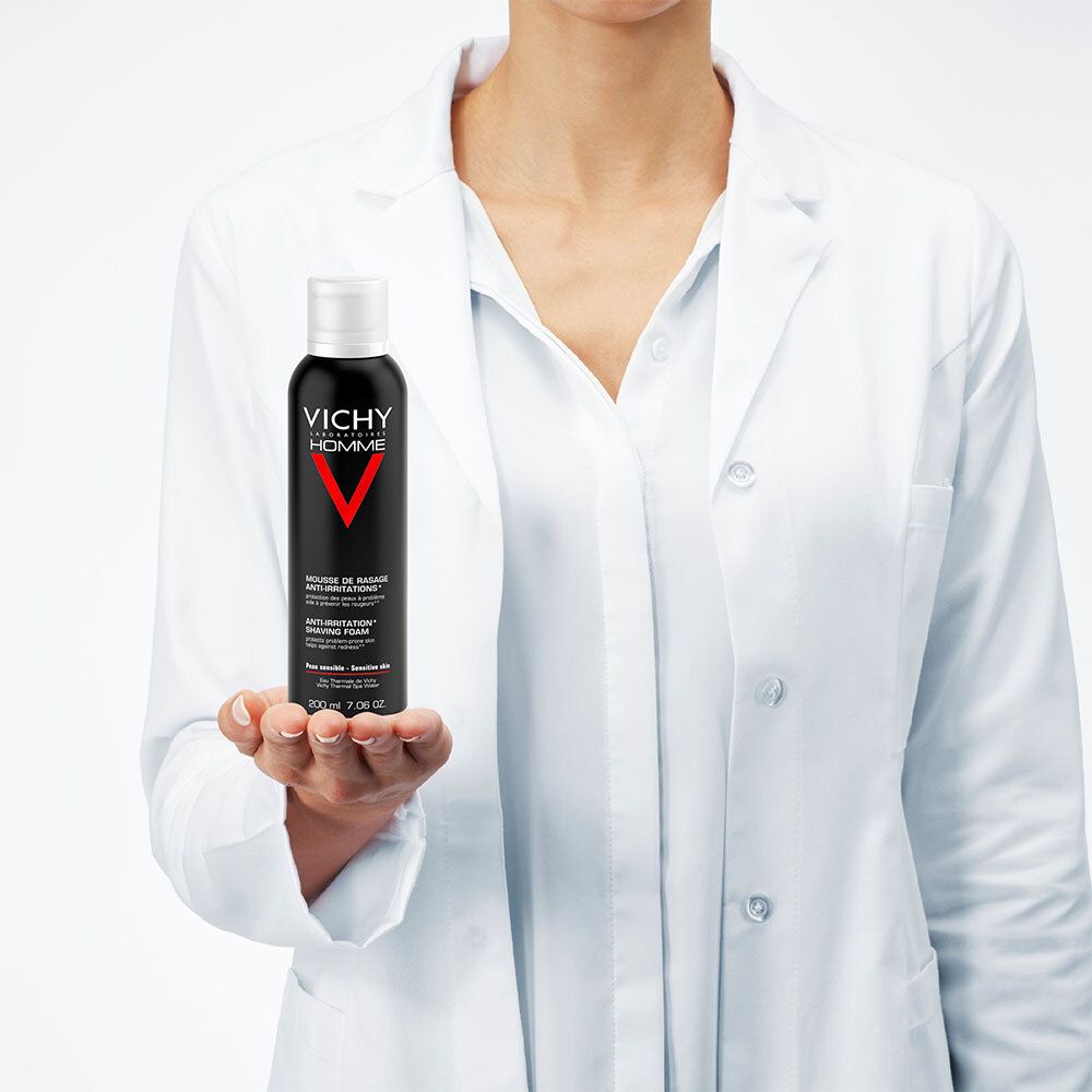Person in weißem Kittel hält eine schwarze Flasche mit weißem Deckel. Aufschrift: VICHY HOMME, rotes V. Text: MOUSSE DE RASAGE ANTI-IRRITATIONS, ANTI-IRRITATION SHAVING FOAM.