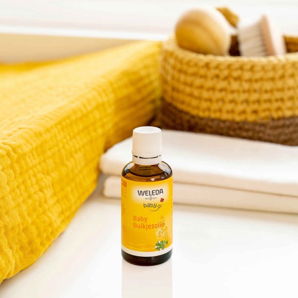 Flacon brun Weleda Baby-Bäuchleinöl. Sur fond jaune. À côté: tissu jaune, panier, serviettes.