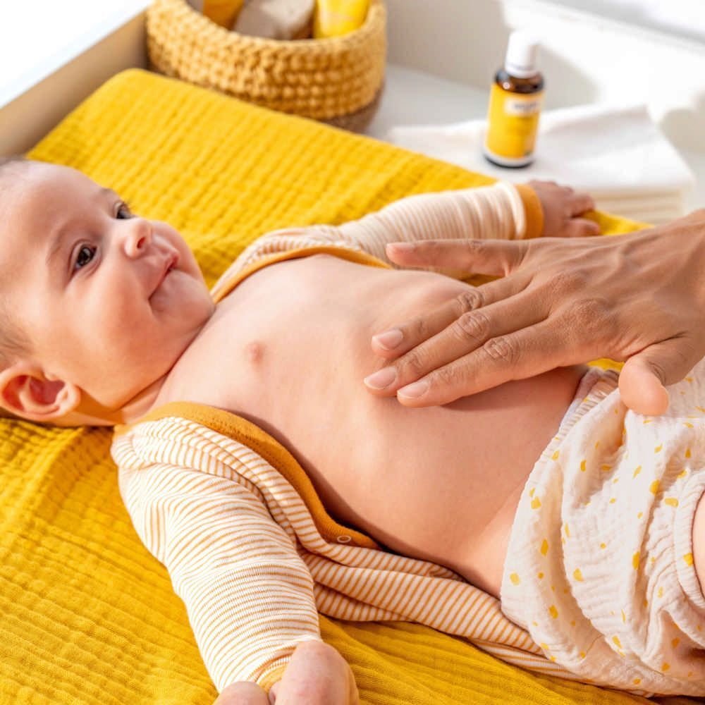 Bébé sur tissu jaune. Main masse le ventre du bébé. Flacon Weleda Baby-Bäuchleinöl hors focus.