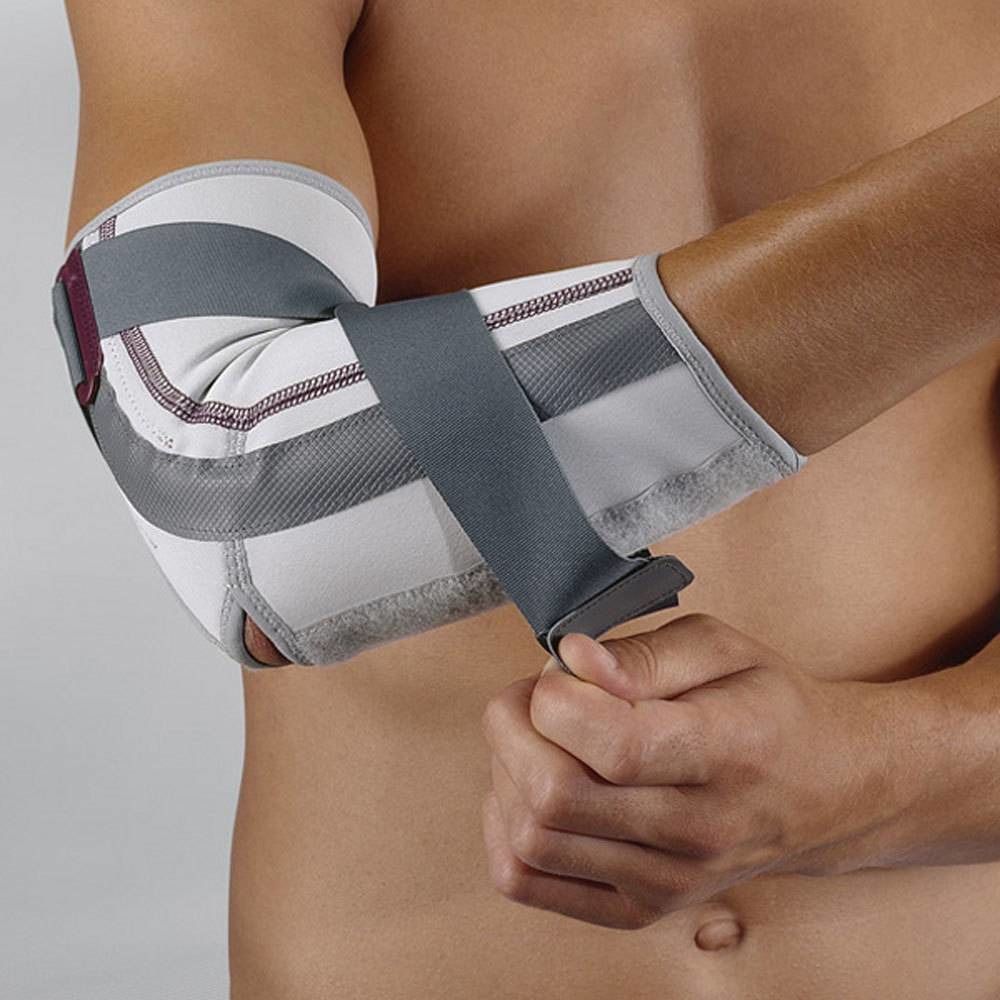 Person trägt eine Ellbogenbandage. Graue Gurte kreuzen sich über dem weißen Stoff. Der Arm ist angewinkelt.