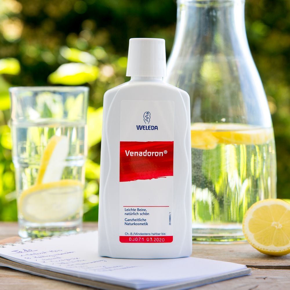 Weleda Venadoron Flasche auf Tisch. Rotes Etikett mit Text. Logo Weleda. Glas Wasser, Zitrone, Karaffe im Hintergrund.