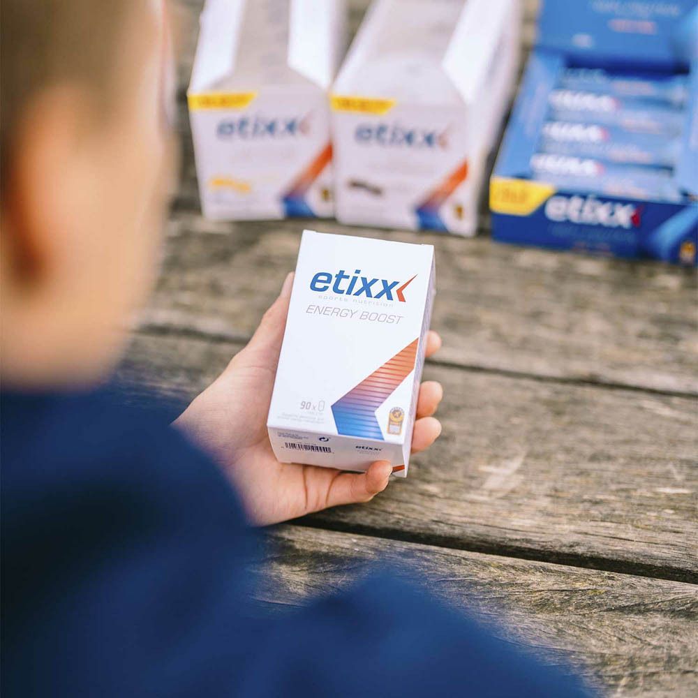 Persoon houdt een witte Etixx-doos vast. Opschrift: Energy Boost. Andere dozen op de achtergrond. Opschrift: Etixx.
