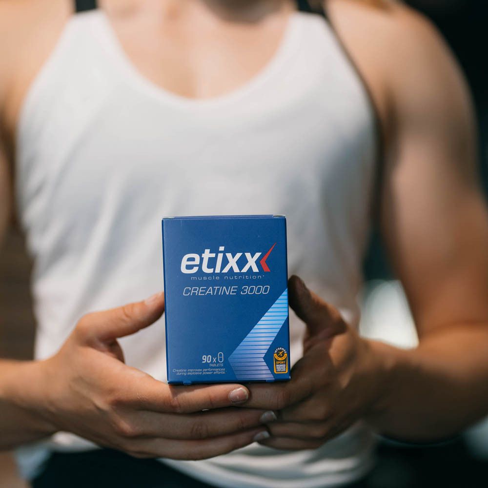 Person hält eine blaue 'etixx Creatine 3000' Packung. Muskeln im Hintergrund sichtbar.