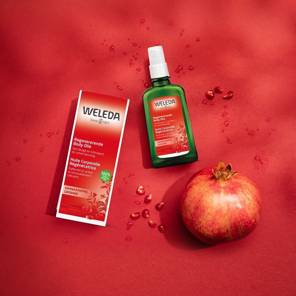 Produktflasche und Verpackung neben Granatapfel auf rotem Hintergrund. Text: Weleda, Regenerierendes Body Oil, Granatapfel.