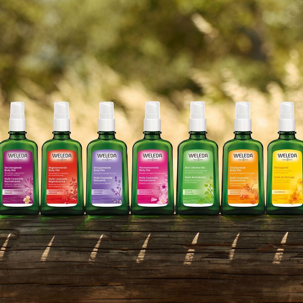 Sieben grüne Flaschen mit farbigen Etiketten. Text: Weleda, Regenerierendes Body Oil. Auf Holzoberfläche.