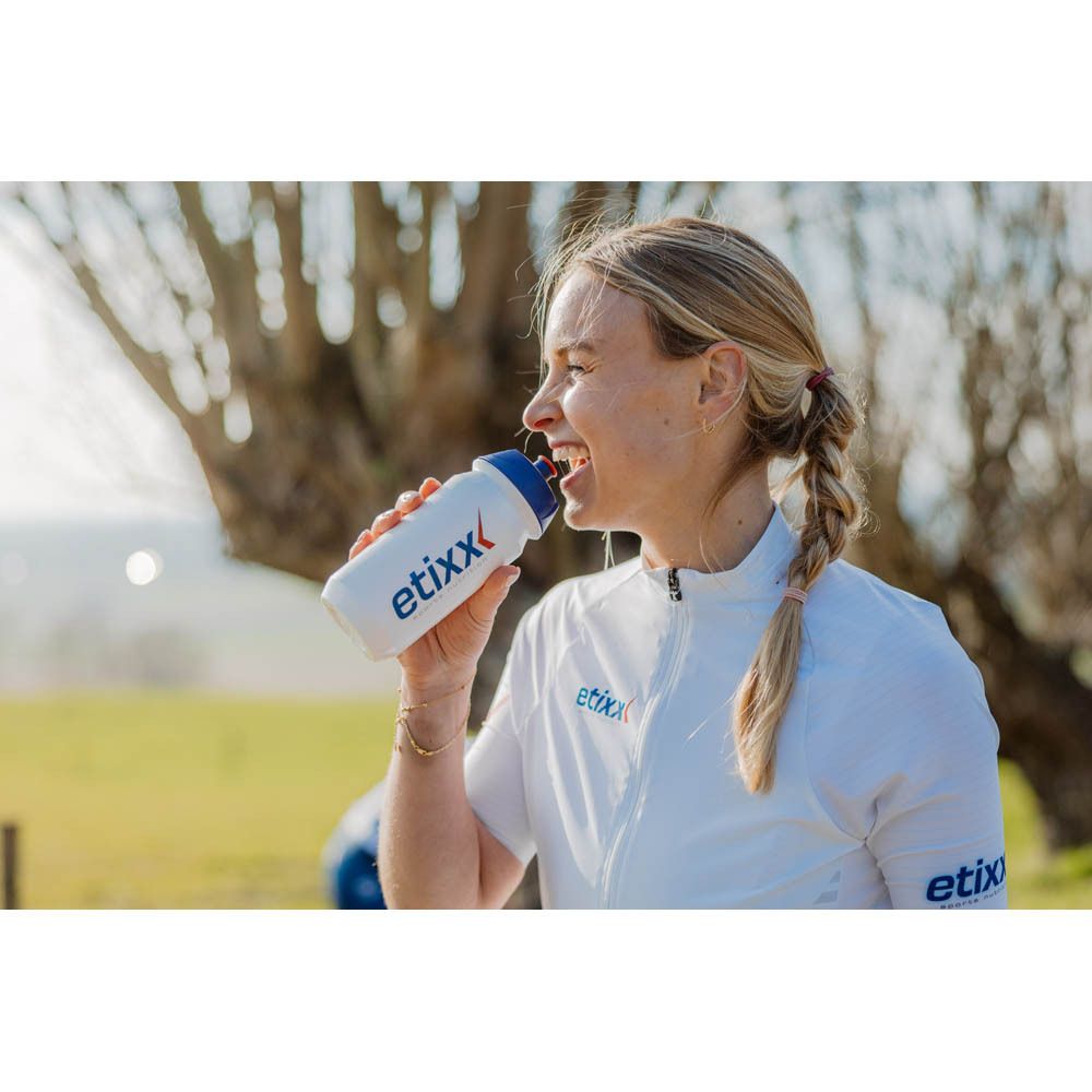 Femme buvant d'une gourde blanche à bouchon bleu. Inscription : etixx. Arrière-plan : arbres.