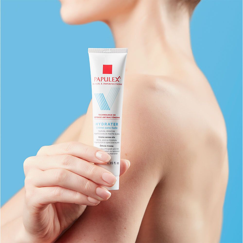 Tube blanc avec carré rouge et logo V bleu. Texte: PAPULEX, HYDRATER Crème sans huile. Tenue par une personne.
