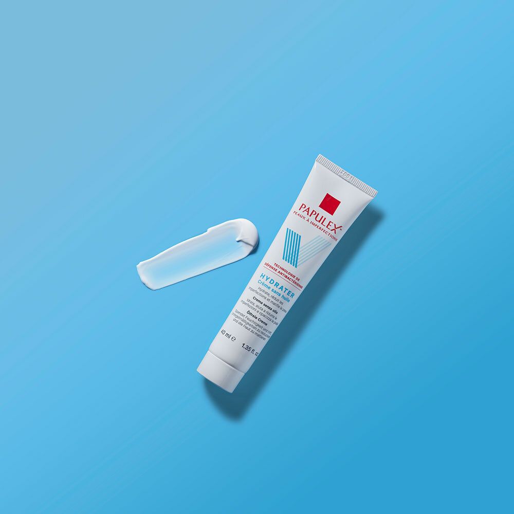 Tube blanc avec carré rouge et logo V bleu. Texte: PAPULEX, HYDRATER Crème sans huile. À côté, une goutte de crème.