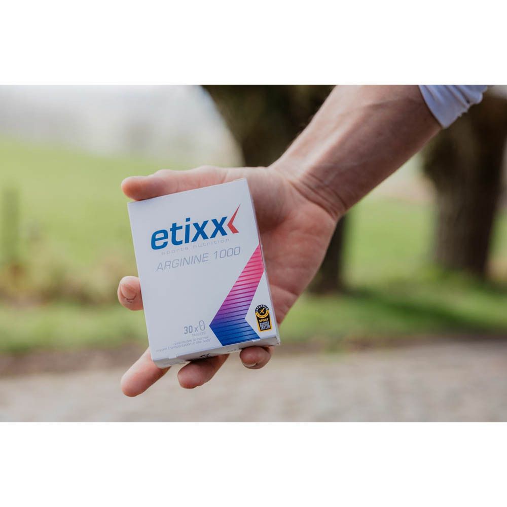Hand houdt een kleine Etixx Arginine 1000-verpakking vast. Witte doos met blauw en rood logo. Vage groene achtergrond.