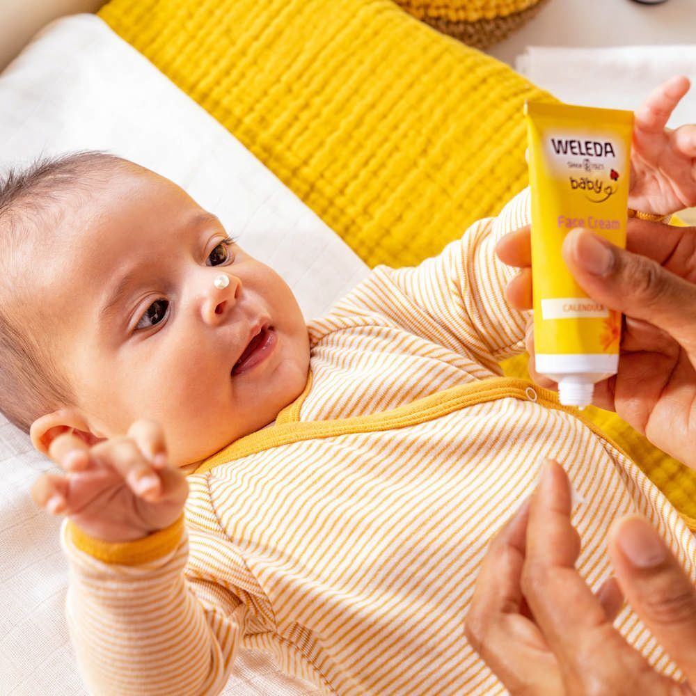 Bébé sur une couverture. Une main tient un tube jaune avec l'inscription WELEDA baby Crème Visage.