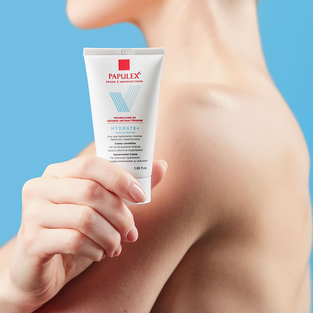 Tube blanc avec logo rouge et bleu. Texte: PAPULEX, Hydrate+ Isocorrection. Tenue par une personne.