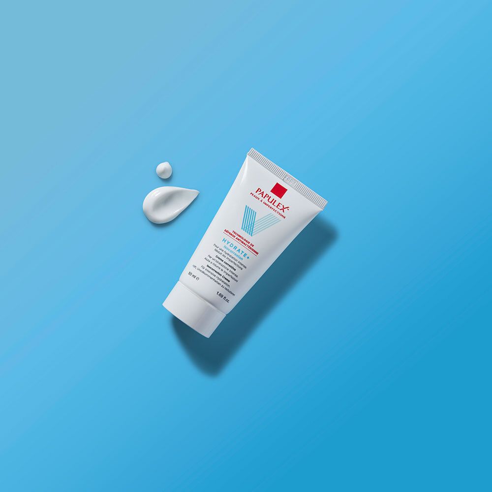 Tube blanc avec logo rouge et bleu. Texte: PAPULEX, Hydrate+ Isocorrection. Deux gouttes de crème à côté.