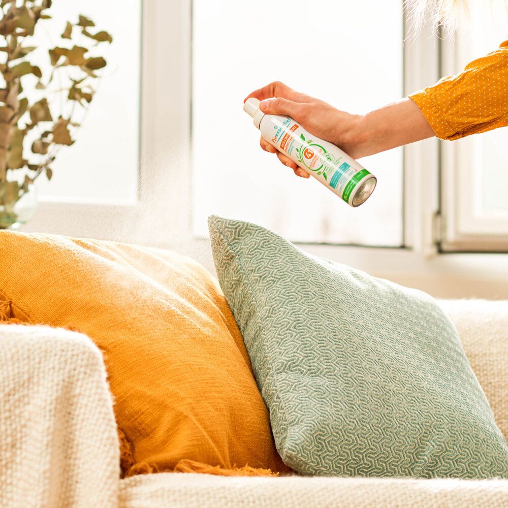 Persoon spuit op kussens. Sprayfles met etiket: Puressentiel, Assainissant.
