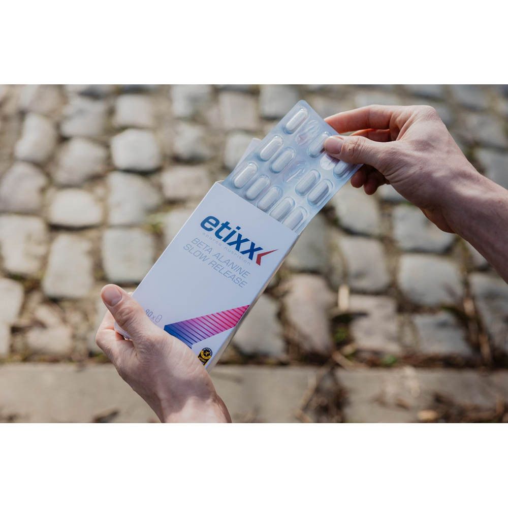 Persoon houdt geopende "etixx" verpakking met tablettenstrips vast. Productnaam "BETA ALANINE SLOW RELEASE" zichtbaar.