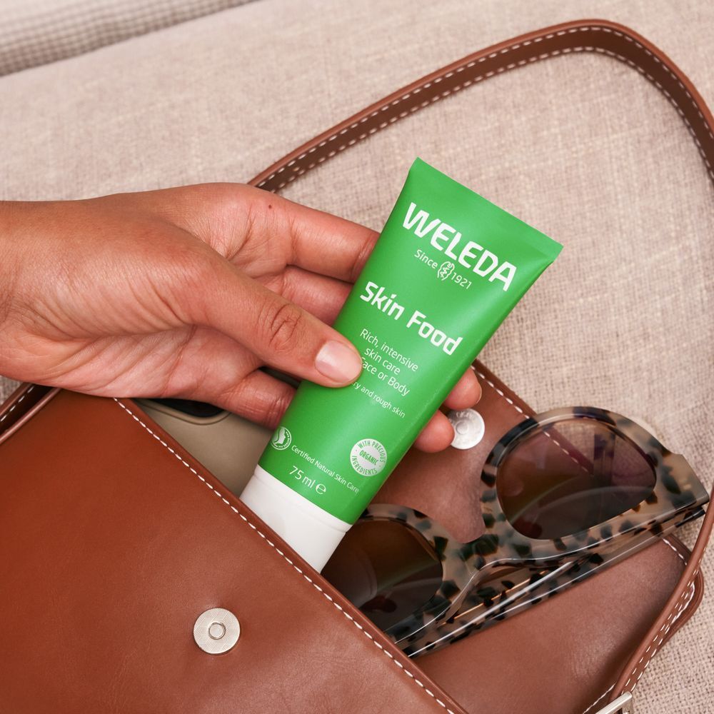 Tube vert Skin Food dans un sac à main marron. Inscription: WELEDA. Des lunettes de soleil sont posées sur le sac.