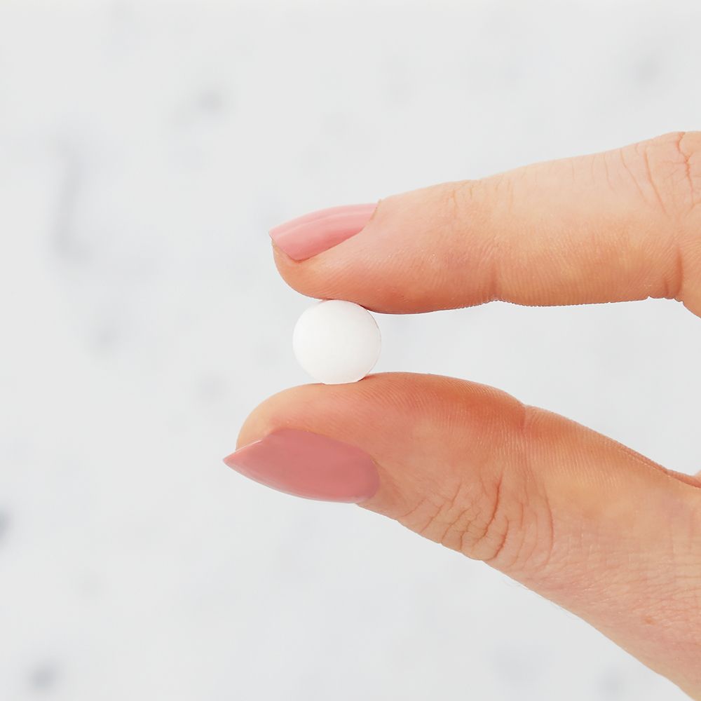 Hand met roze nagels die een witte tablet tussen duim en wijsvinger vasthoudt.