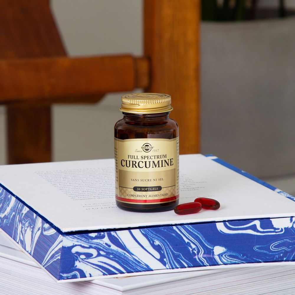 Bruine fles met gouden dop en twee rode capsules op een boek. "Full Spectrum Curcumin" etiket zichtbaar.