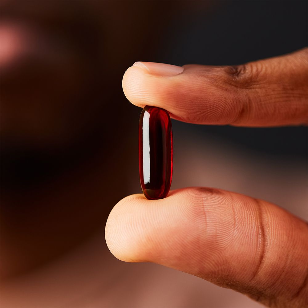 Een rode, langwerpige capsule tussen twee vingers. Donkere achtergrond.