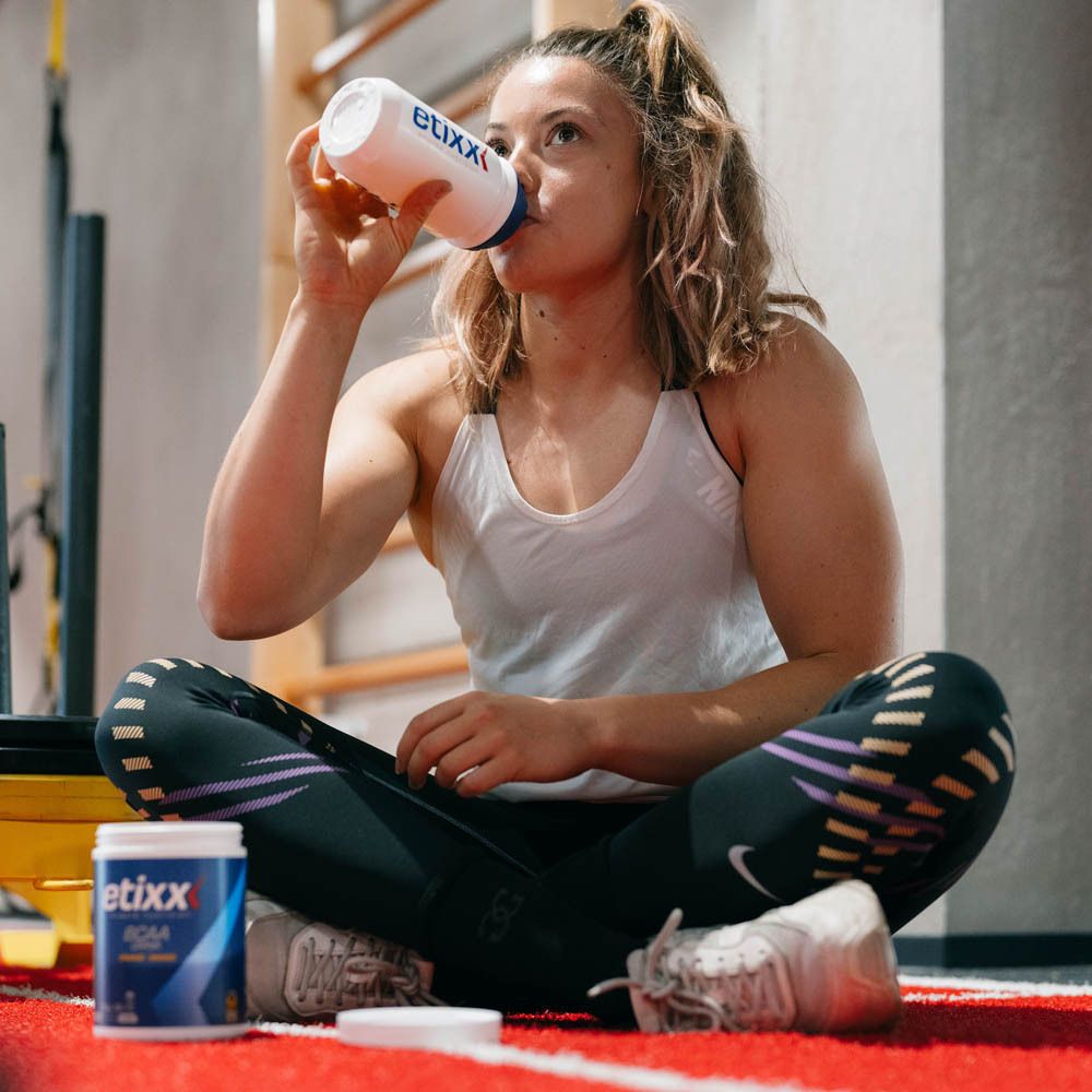 Athletin trinkt aus einer weißen Flasche. Im Hintergrund Sportgeräte. Dose mit etixx BCAA Drink.
