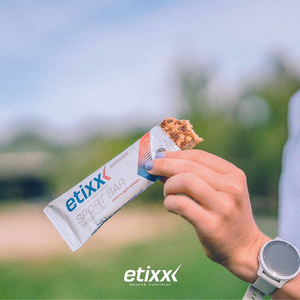 Personne tenant une barre énergétique Etixx à moitié mangée. Inscription: Natural Oat, Sweet & Salty Caramel. Montre au poignet.