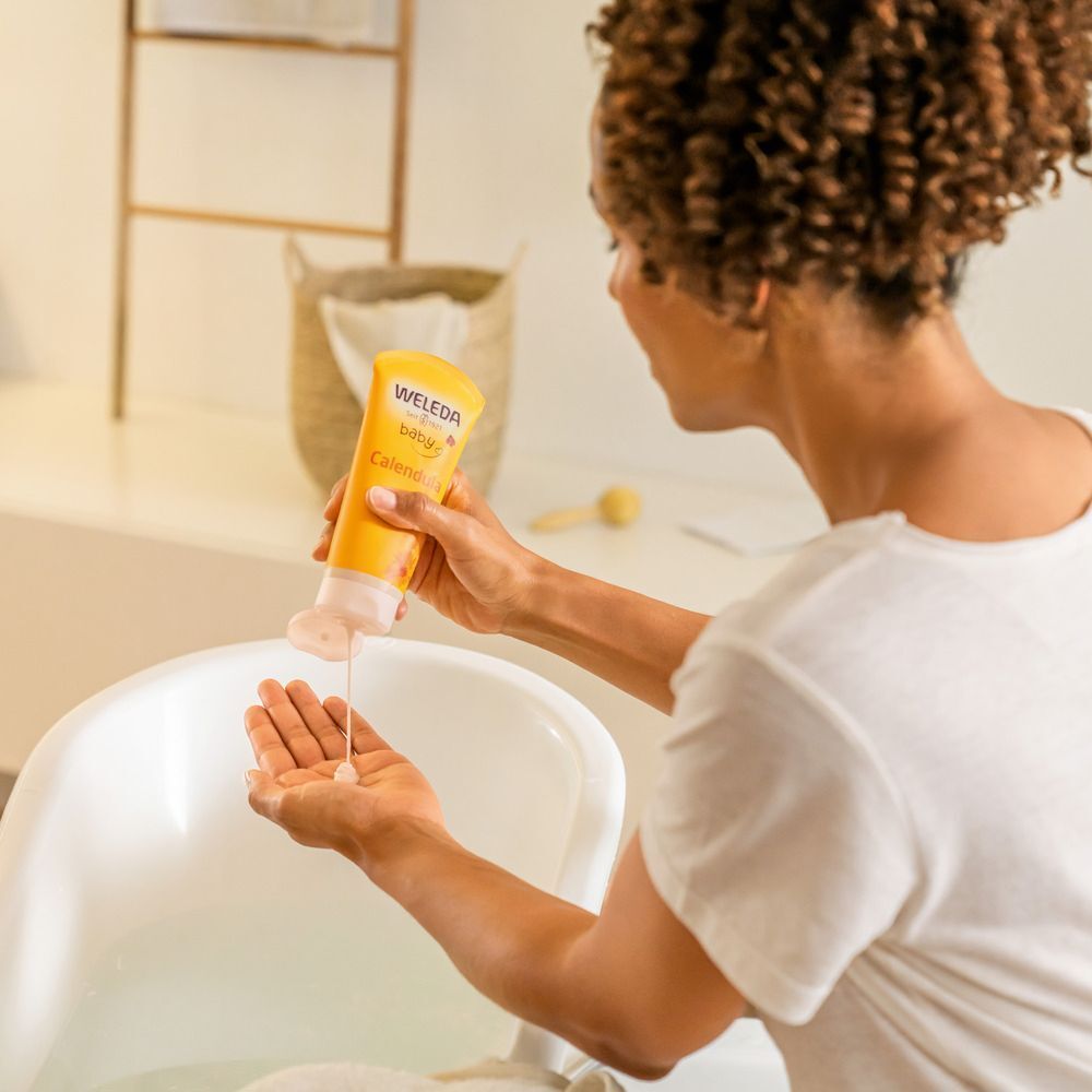 Vrouw houdt gele tube "Weleda Baby" vast en giet product in haar hand. Product: Shampoo & Duschcreme. Badkuip op de achtergrond.