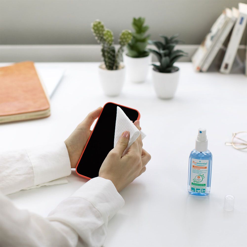 Persoon reinigt smartphone met doek. Naast, blauwe fles met verstuiver.