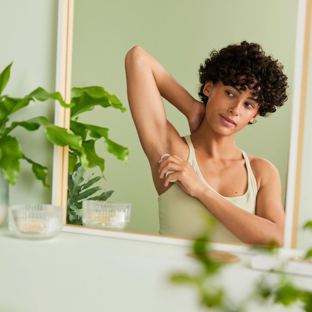 Femme dans miroir, roll-on déodorant sous le bras. Miroir, mur vert, plantes, environnement clair.