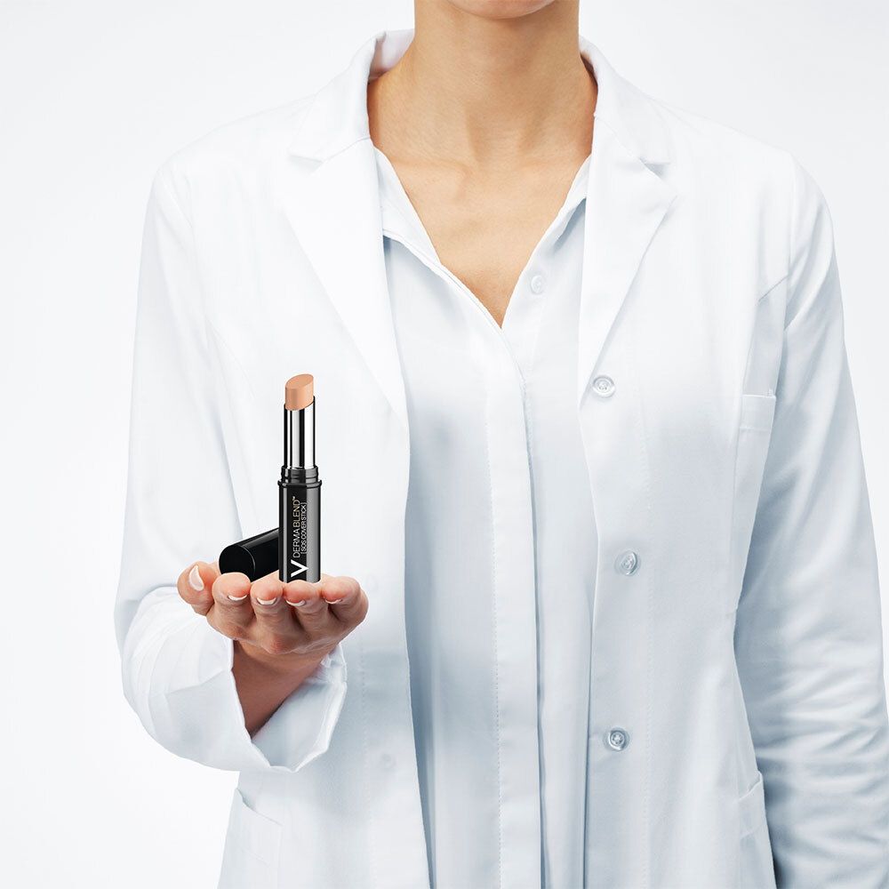 Persoon in witte jas houdt concealer stick vast. Merk en naam zichtbaar.