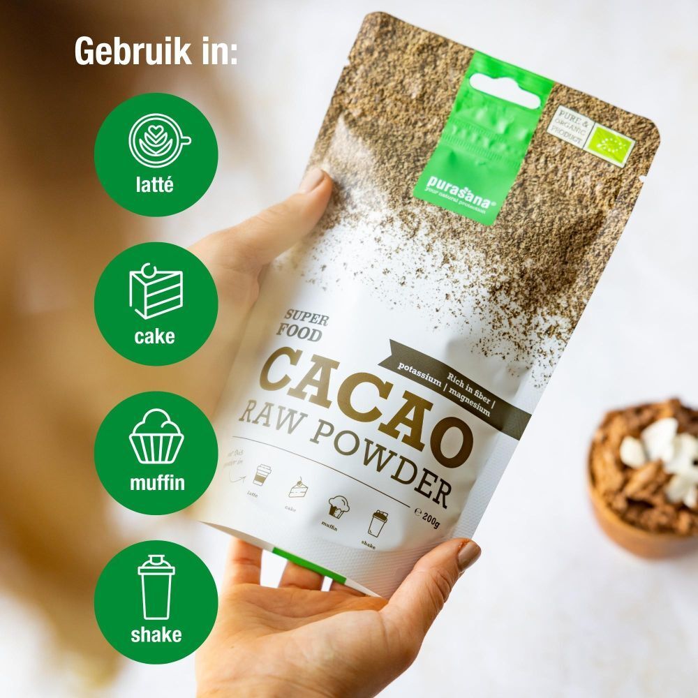 Hand houdt zakje purasana® CACAO RAW POWDER. Tekst: Gebruik in: Latté, Cake, Muffin, Shake. Groen en wit.