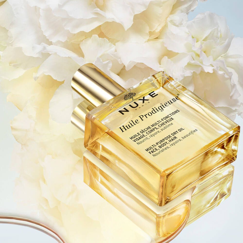 NUXE Huile Prodigieuse® Trockenöl-Flasche. Liegend, auf spiegelnder Oberfläche. Blüten im Hintergrund.