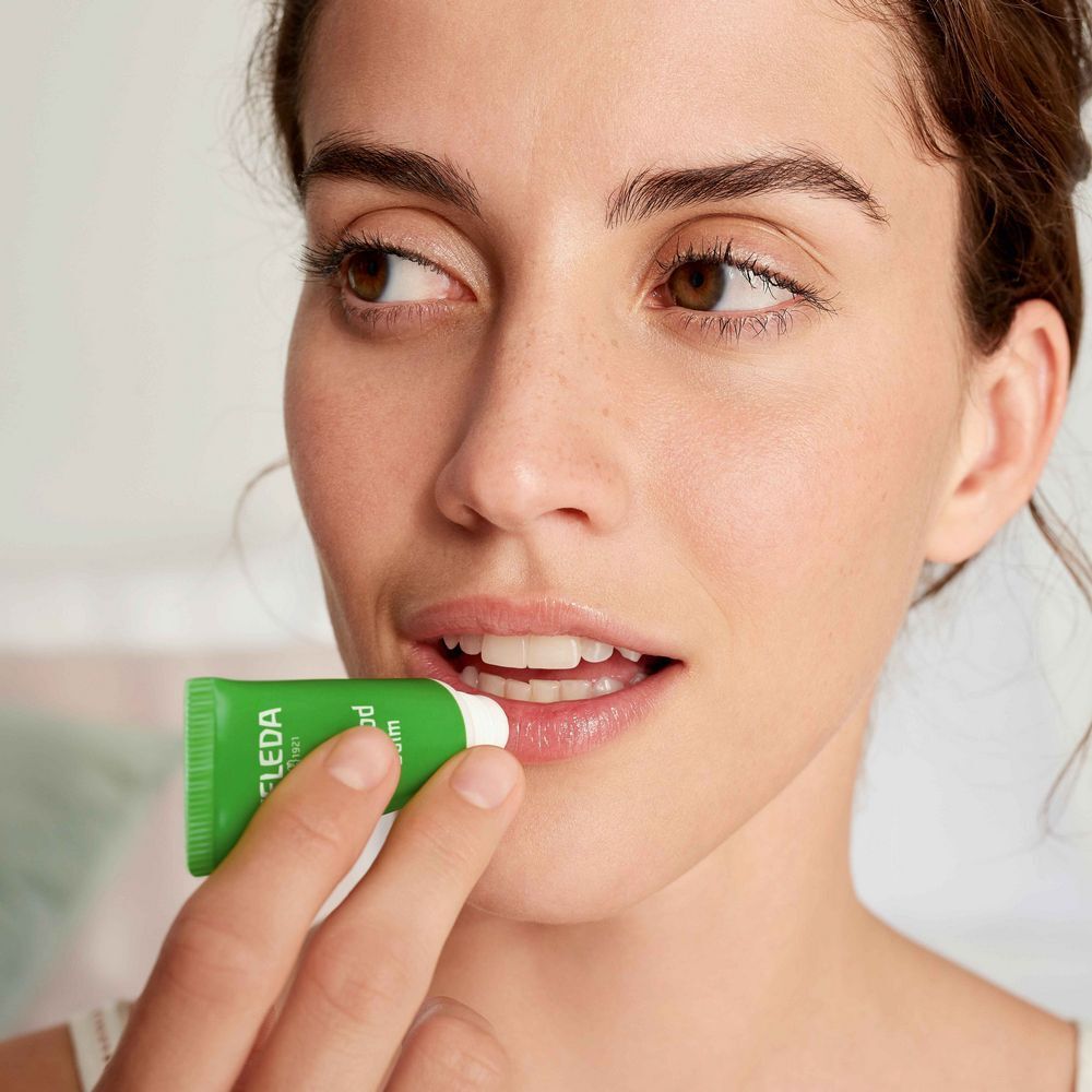 Frau trägt Lippenbalsam auf. Grüne Tube mit weißem Verschluss. Aufschrift: Weleda Skin Food.