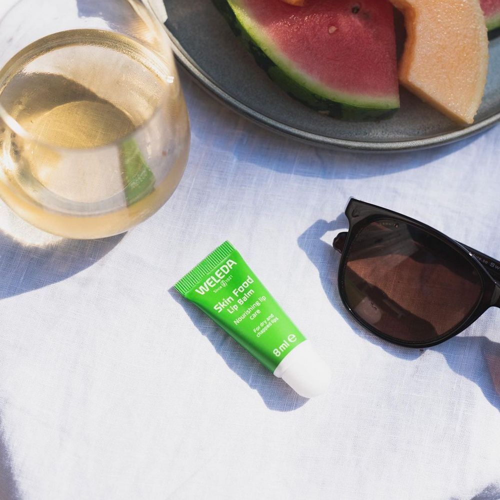 Lippenbalsam neben Sonnenbrille, Glas und Obst. Grüne Tube mit weißem Verschluss. Aufschrift: Weleda Skin Food Lip Balm.