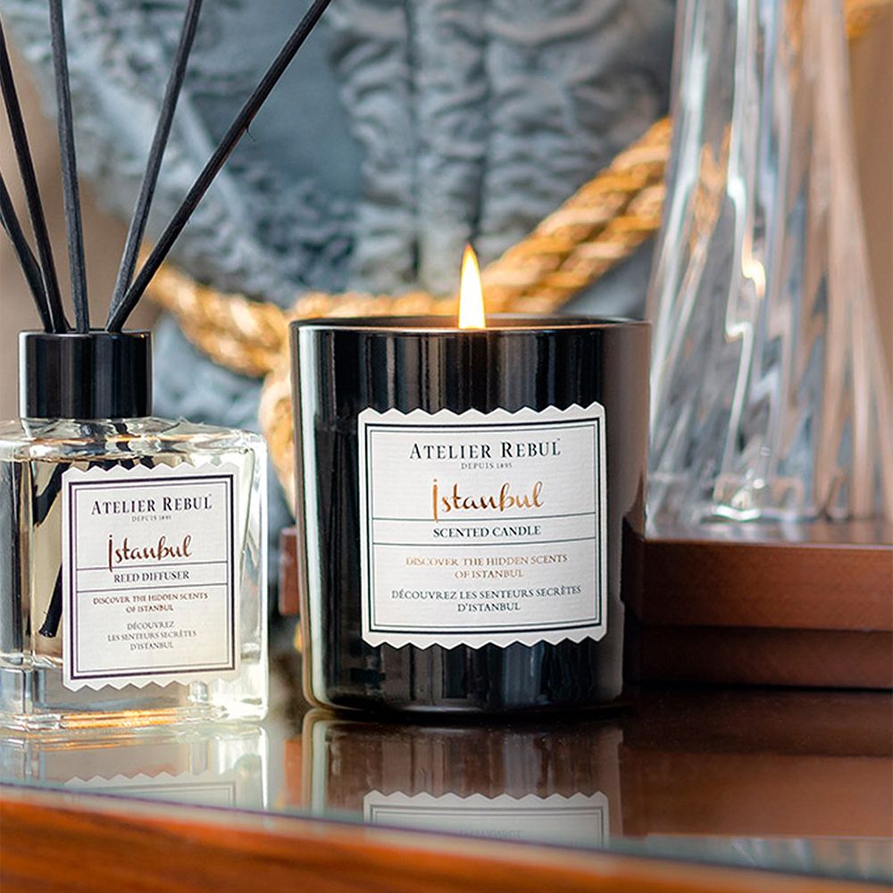 Bougie allumée. Verre noir, étiquette: ATELIER REBUL, Istanbul, Scented Candle. À côté d'un diffuseur.