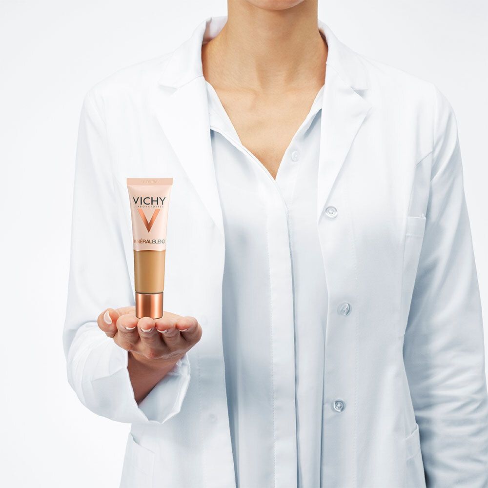 Personne tenant le fond de teint. Marque: Vichy. Tube avec contenu brun. La personne porte une blouse blanche.