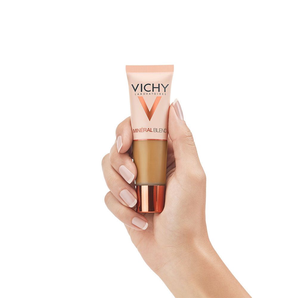 Main tenant le fond de teint. Marque: Vichy. Tube avec contenu brun. Tube beige avec bouchon cuivré.