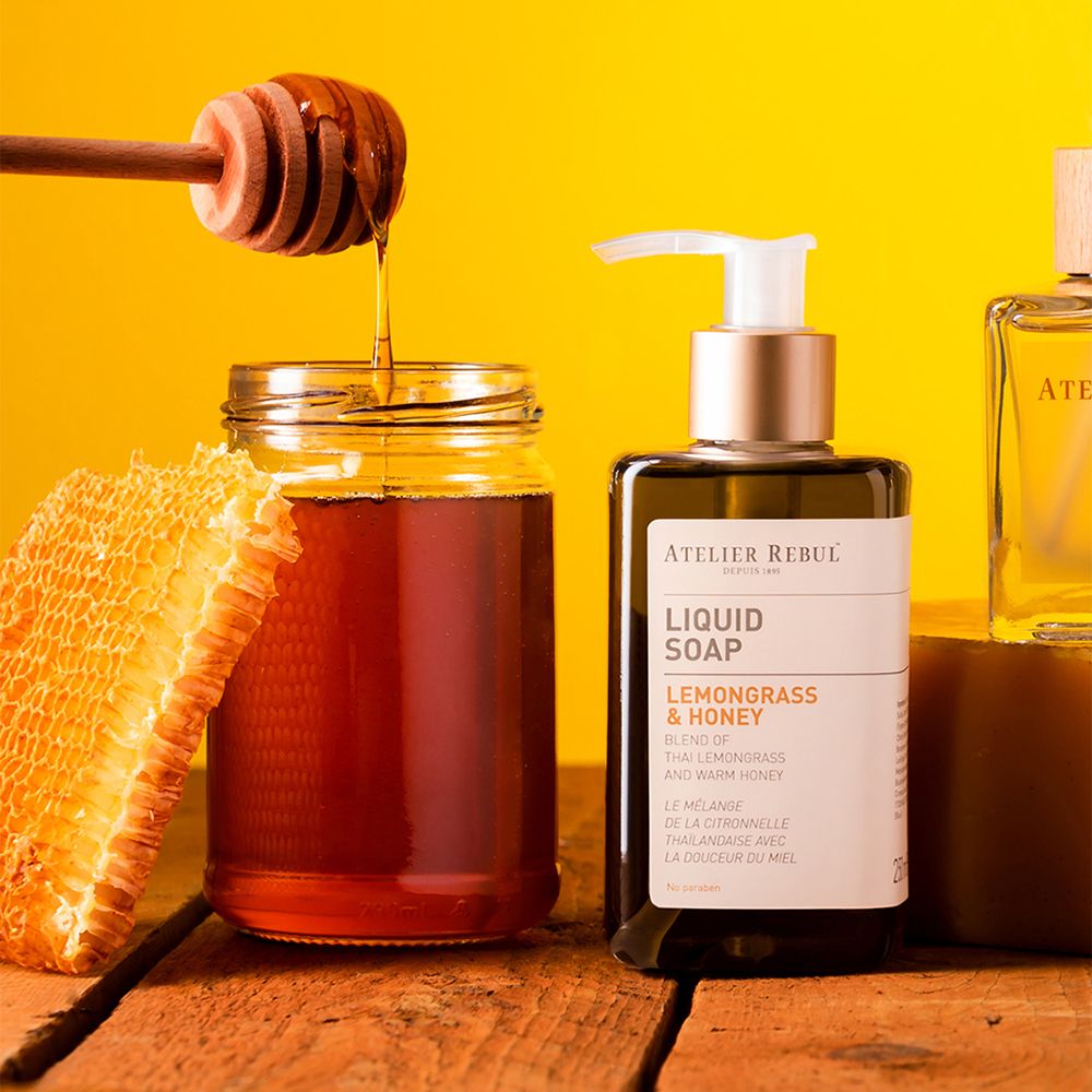 Pot honing, honingraat en honingdispenser. Opschrift: ATELIER REBUL, LIQUID SOAP, LEMONGRASS & HONEY. Gele achtergrond.