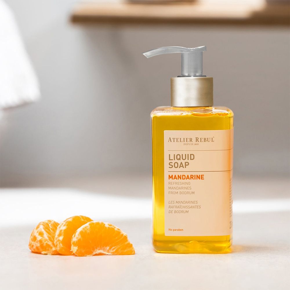 Vloeibare zeep met pomp en mandarijnpartjes. Transparante houder met oranje vloeistof. Opschrift: ATELIER REBUL, LIQUID SOAP.