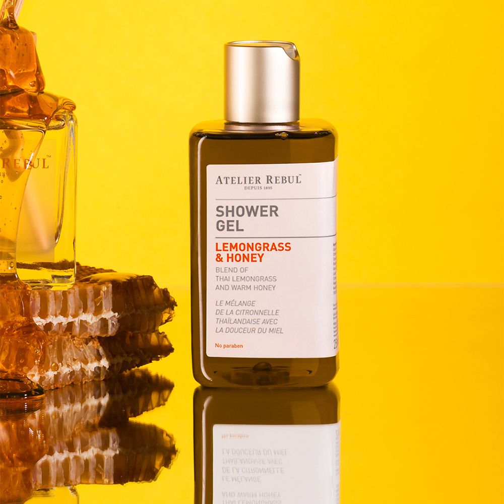 Bruine fles met gouden dop. Opschrift: Shower Gel, Lemongrass & Honey. Gele achtergrond.