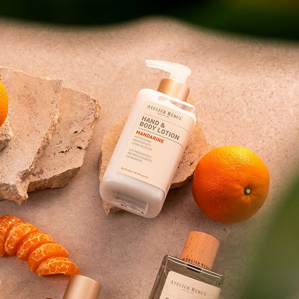 Hand- und Bodylotion-Spender neben Mandarinen und einem Parfümflakon. Aufschrift: ATELIER REBUL, Hand & Body Lotion, Mandarine.