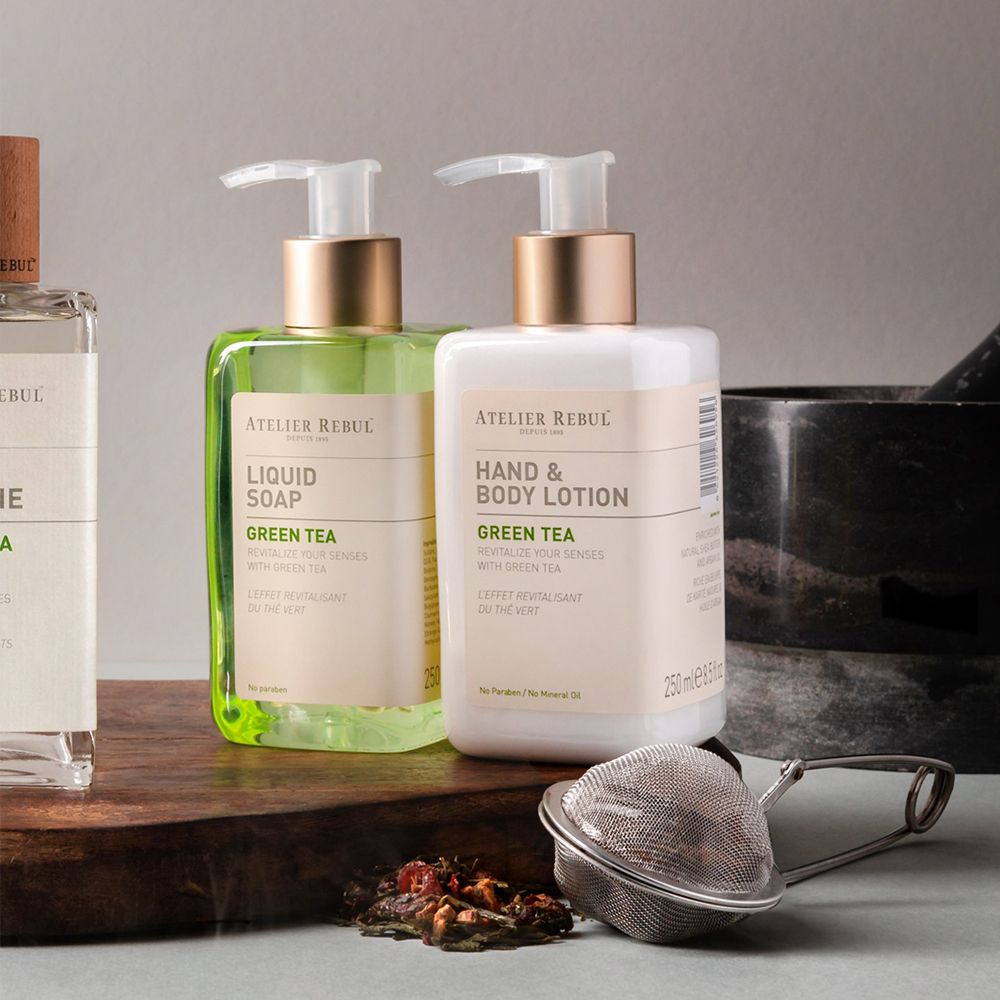 Zwei Flaschen Hand- und Bodylotion und Flüssigseife. Aufschrift: ATELIER REBUL, Green Tea. Dazu ein Teesieb und Teeblätter.