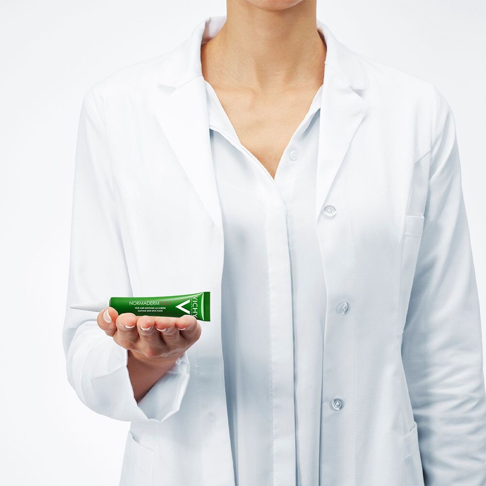 Personne tenant un tube vert. Nom du produit : Normaderm. La personne porte une blouse blanche.