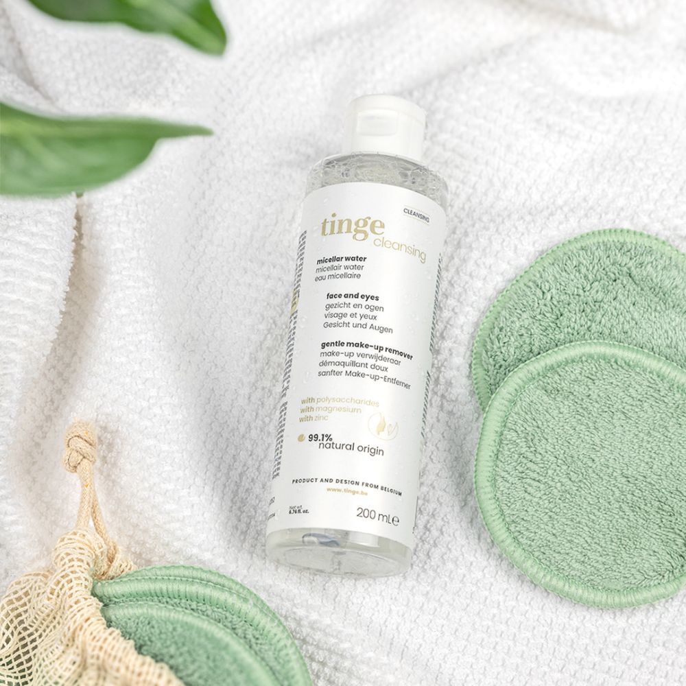 Fles op witte handdoek, naast groene make-up remover pads. Label "tinge cleansing micellair water".