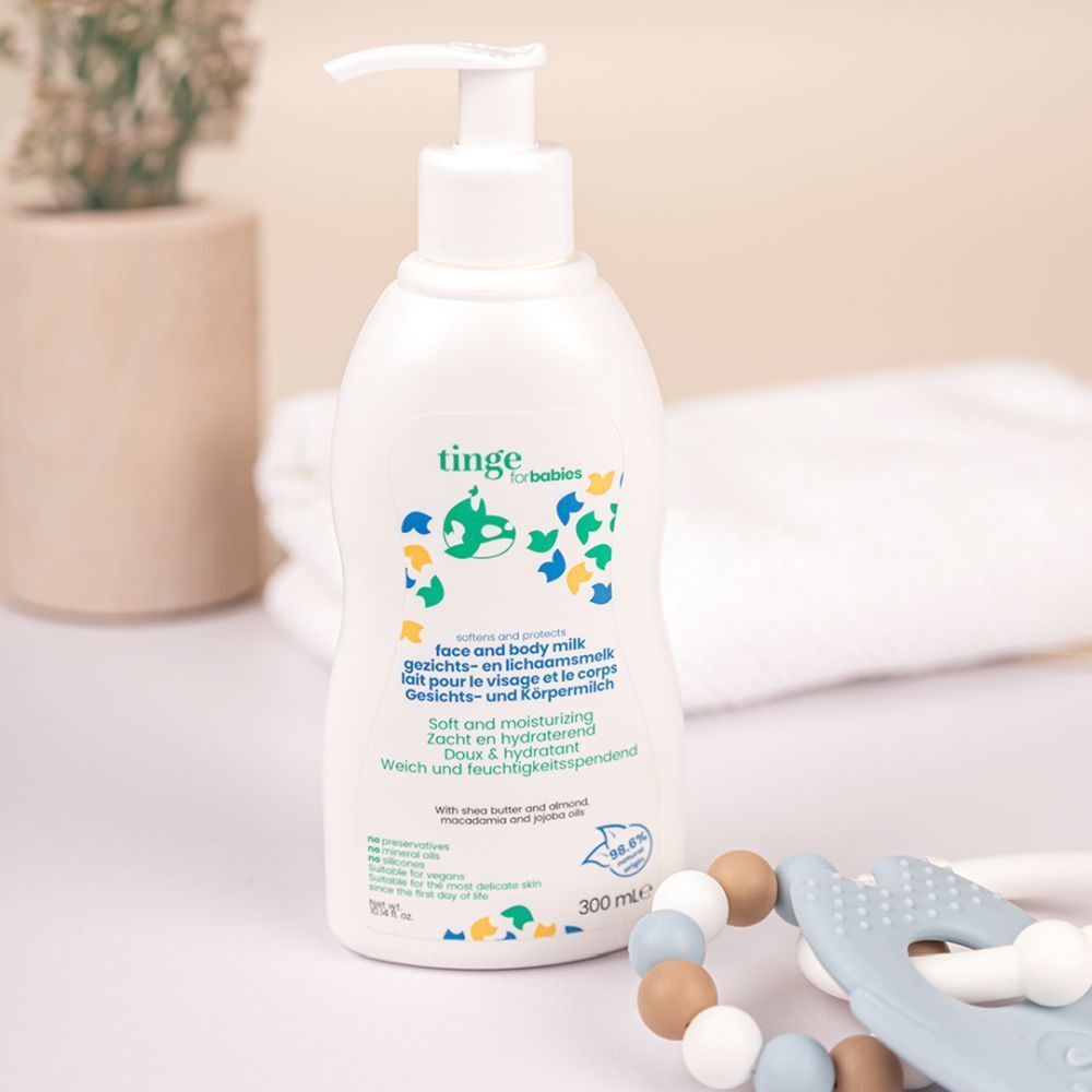 Witte fles met pomp. Opschrift: tinge for babies, gezichts- & lichaamsmelk. 300 ml. Achtergrond: textiel en bijtring.